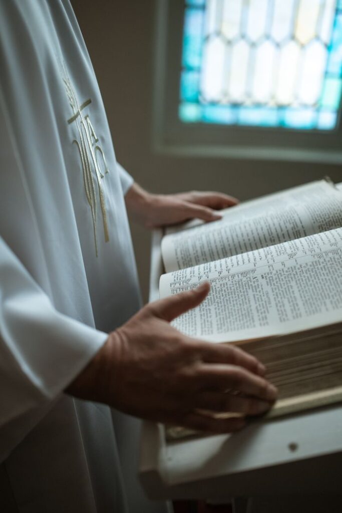 Close-up of a priest holding an open Bible indoors with stained glass window behind.